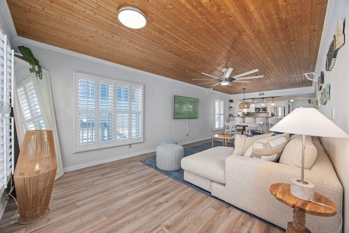 A spacious living area with a wooden ceiling, a large window with blinds, a sofa, a table with a lamp, and a kitchen in the background.