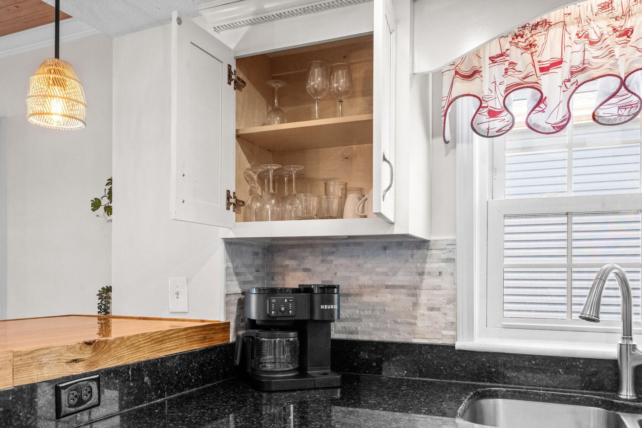 Una cocina con cafetera integrada, un aparador para copas de vino y una ventana con cortinas.