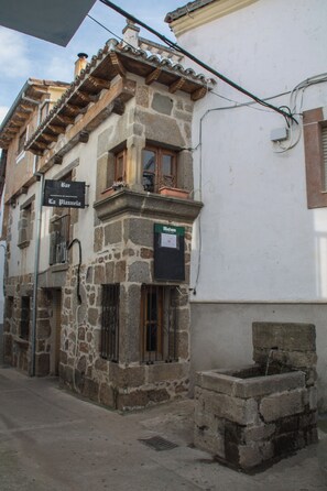 Un bâtiment en pierre avec une enseigne de bar, une fenêtre ornée d’une grille métallique et une petite fontaine extérieure.