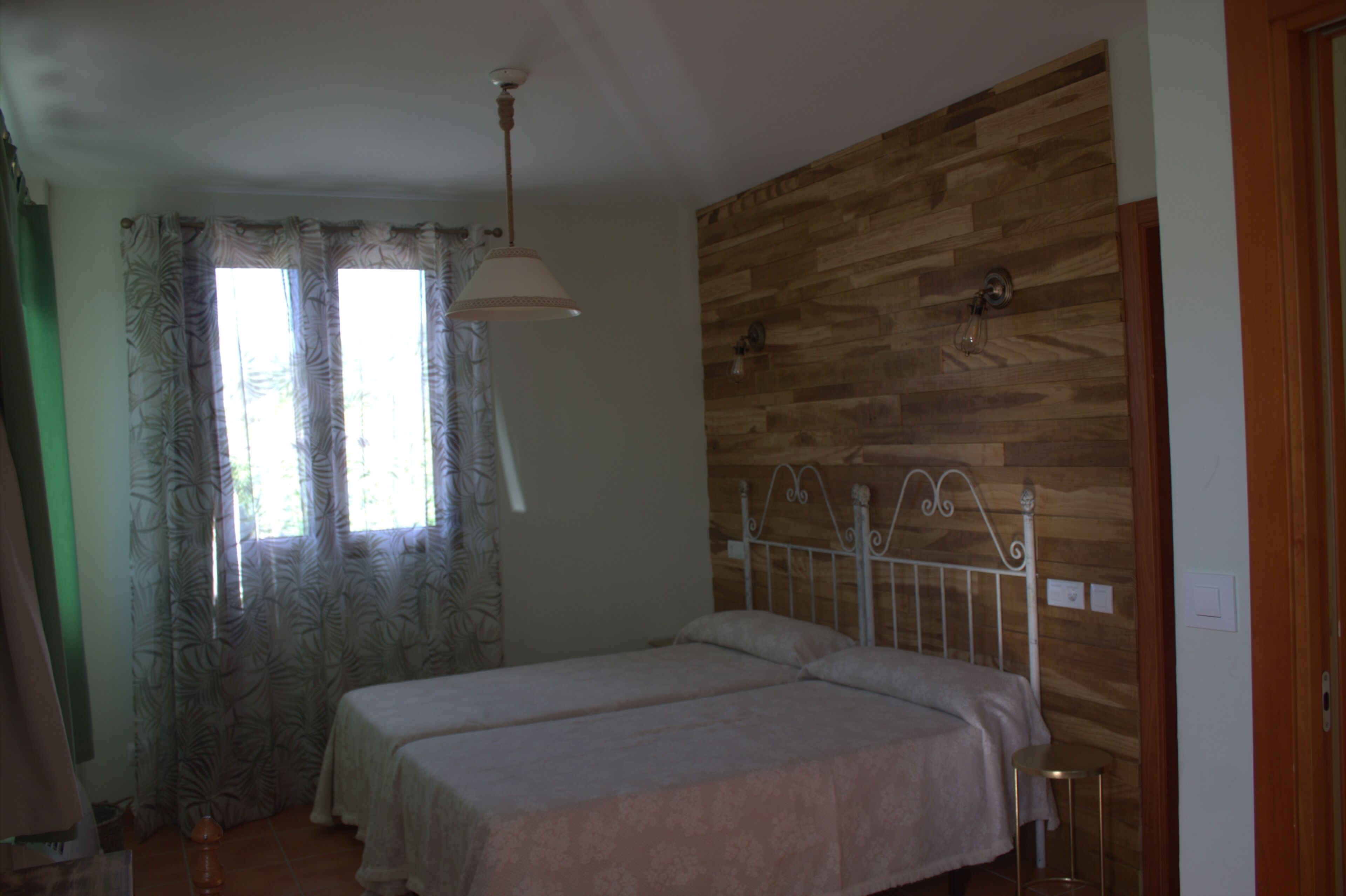 A bedroom with two beds, a wooden wall, a window with curtains, and a hanging light fixture.