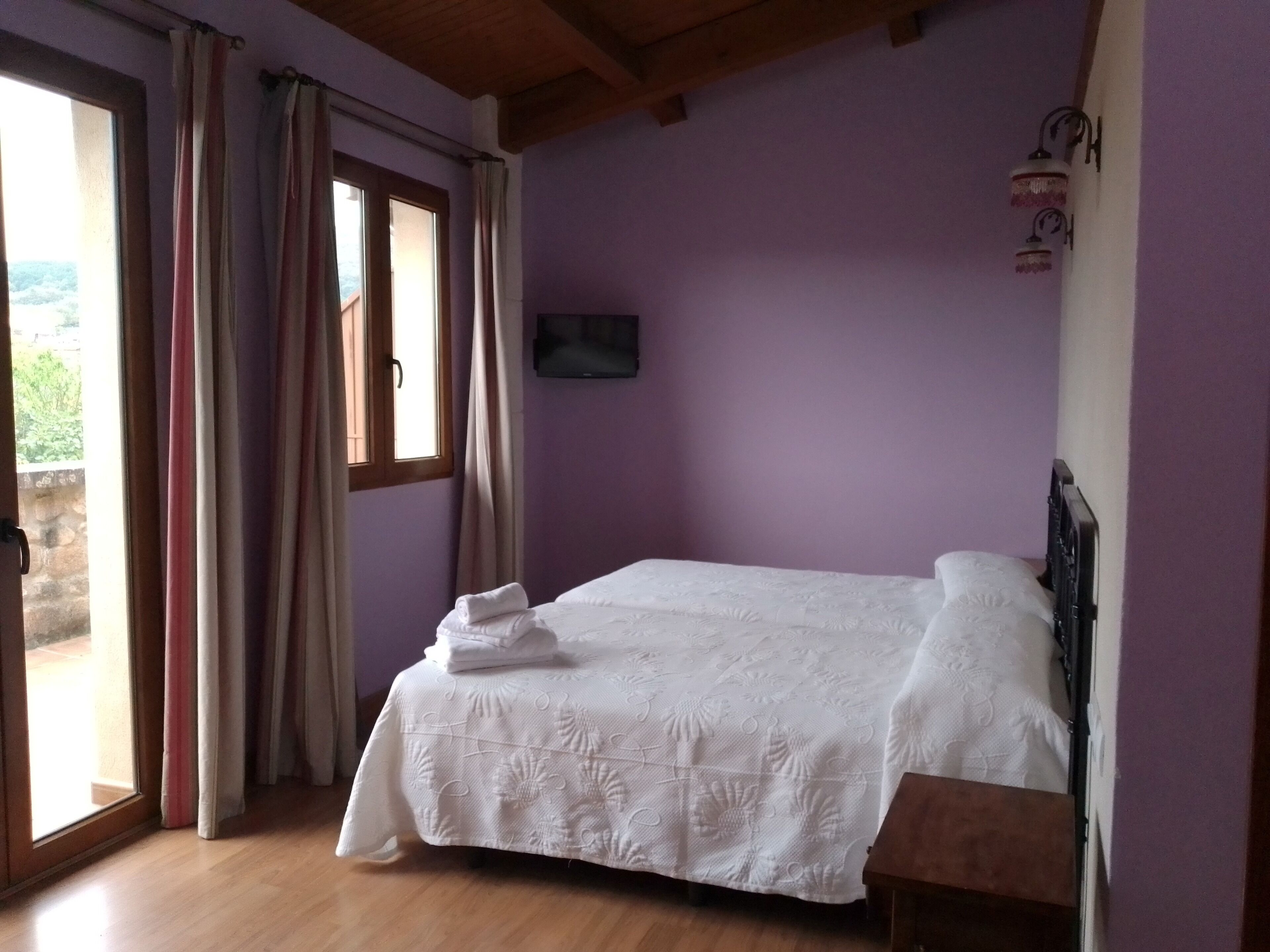 Une chambre à coucher avec un lit, un plancher en bois, une fenêtre avec des rideaux et un téléviseur fixé au mur.