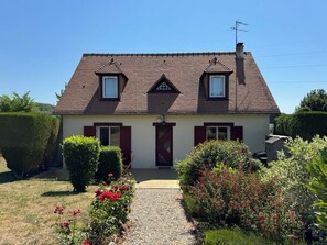 A two-story house with a sloped roof, a front entrance, and a small garden with flowers and shrubs. - Quiet house with garden, 5 min from Deauville (Touques)