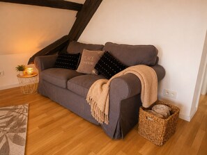Ein graues Sofa mit einer Tagesdecke, ein kleiner Beistelltisch mit Pflanze und Kerze sowie ein gewebter Korb mit Handtüchern.