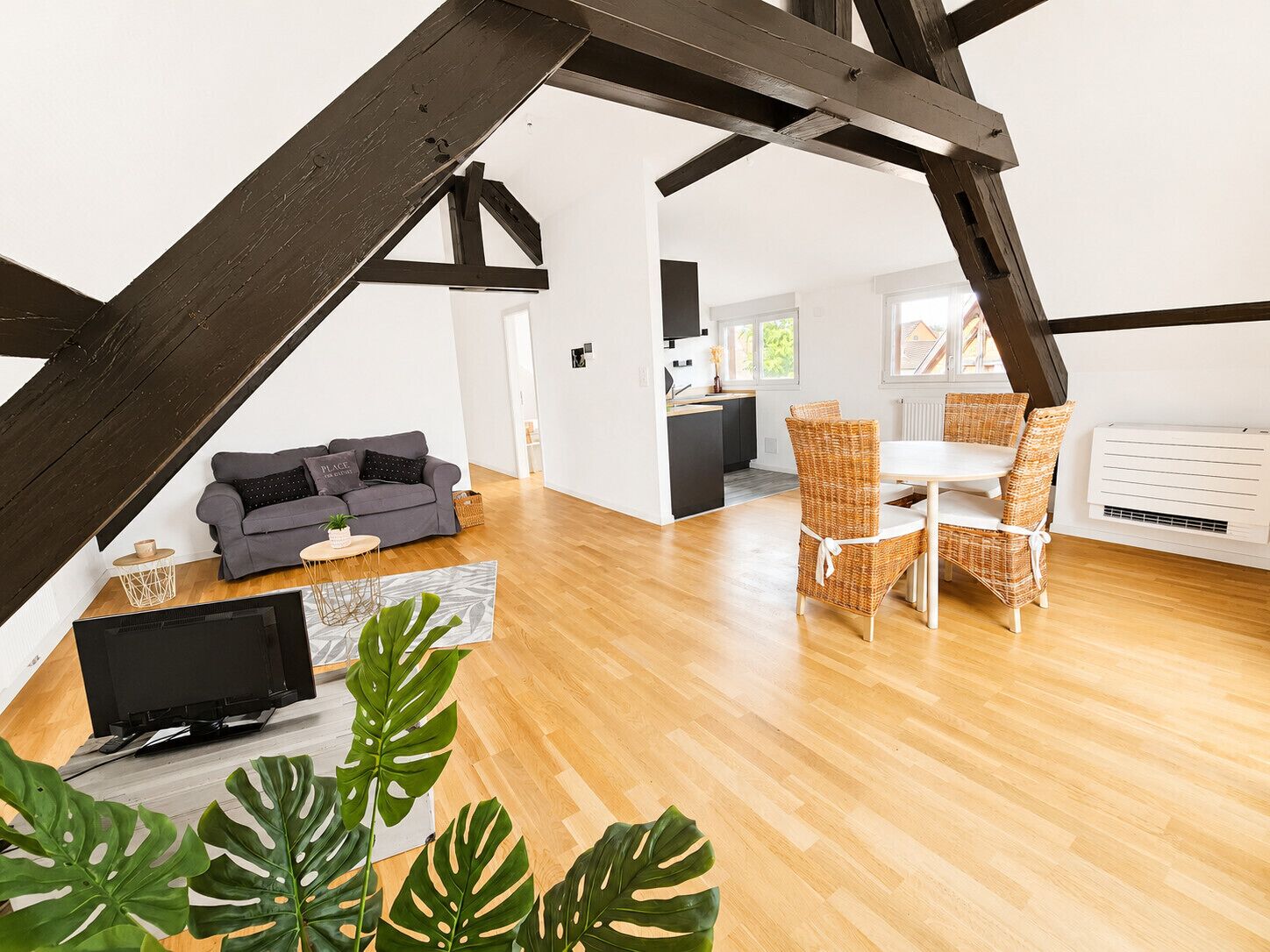 A modern loft with exposed wooden beams, a grey sofa, wicker dining chairs, and a flat-screen TV.