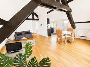 A modern loft with exposed wooden beams, a grey sofa, wicker dining chairs, and a flat-screen TV. - A Cozy Nest Under the Roof (Wiwersheim)