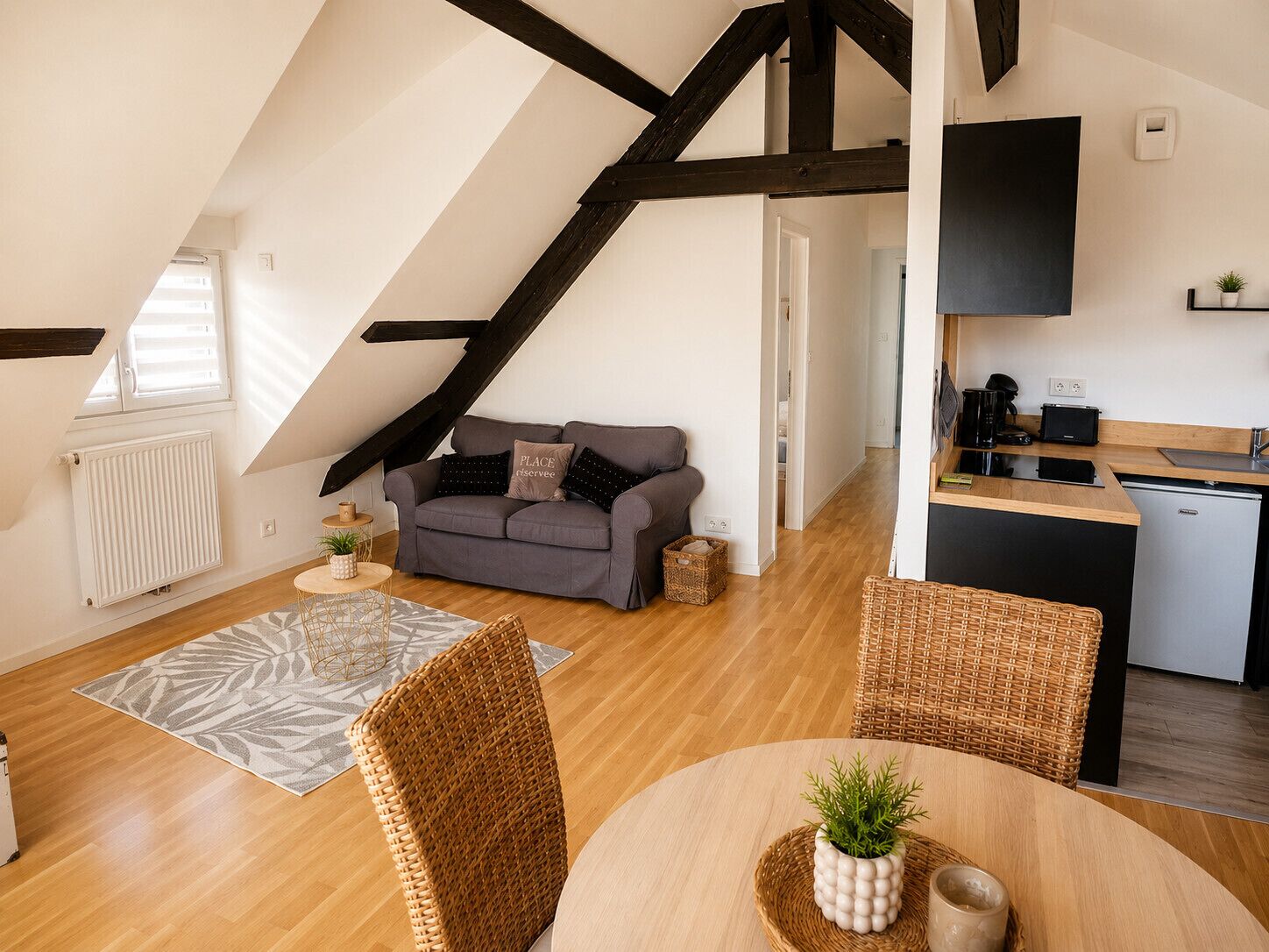 Un appartement moderne de style « loft » comprenant un lit en mezzanine, un coin cuisine, un espace repas avec une table ronde et un canapé gris agrémenté de coussins décoratifs.