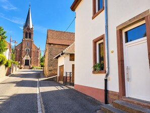 A street leading to a church with a bell tower, a white building with a door, and a window with a plant. - A Cozy Nest Under the Roof (Wiwersheim)