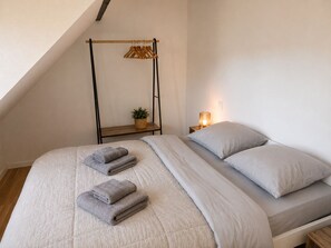 Ein Schlafzimmer mit einem Bett, Kissen, einem Nachttisch mit Lampe, einem hängenden Holzgestell und einer kleinen Pflanze.