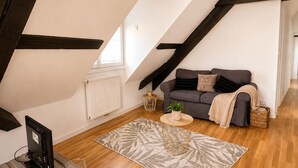 A loft-style room with a sloped ceiling, a grey sofa with a blanket, a small round table, a radiator, and a wooden floor. - A Cozy Nest Under the Roof (Wiwersheim)