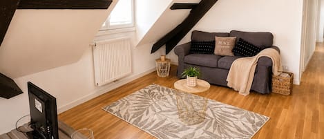 Habitación estilo loft con techo inclinado, un sofá gris con una manta, una mesita redonda pequeña, un radiador y suelo de madera.