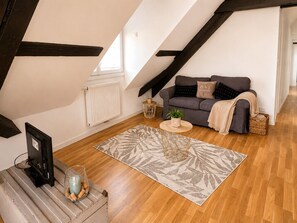 A loft-style room with a sloped ceiling, a grey sofa with a blanket, a small round table, a radiator, and a wooden floor. - A Cozy Nest Under the Roof (Wiwersheim)