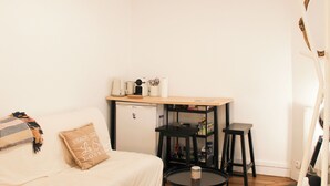 En kompakt opholdsstue med sofa, lille køleskab, kaffemaskine, bord og bogreol.