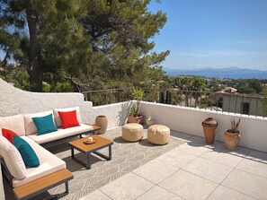 Une terrasse extérieure baignée de soleil, avec un canapé blanc, des poufs et une table en bois.