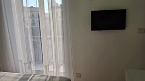 Een kamer met een bed, een aan de muur bevestigde televisie en een raam met dunne gordijnen.