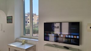 Ein modernes Wohnzimmer mit einem großen Flachbildfernseher an der Wand, einem kleinen Tisch mit einer Fernbedienung und einem Fenster mit Blick auf Gebäude.