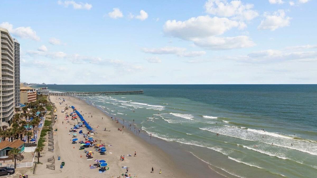 A beach with a pier extending into the ocean, a hotel with balconies overlooking the beach, and umbrellas and beachgoers.
