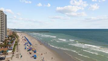 A beach with a pier extending into the ocean, a hotel with balconies overlooking the beach, and umbrellas and beachgoers.