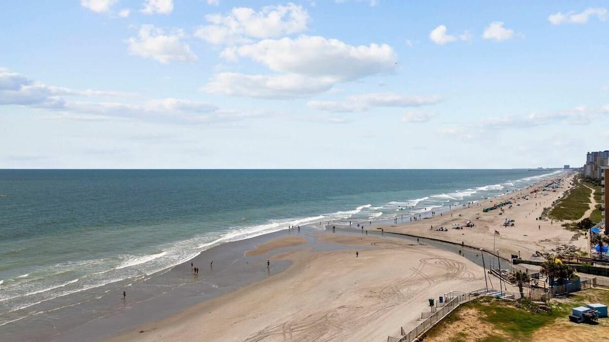 En strand med en bugtende vej, sandet bredde og bygninger i baggrunden.