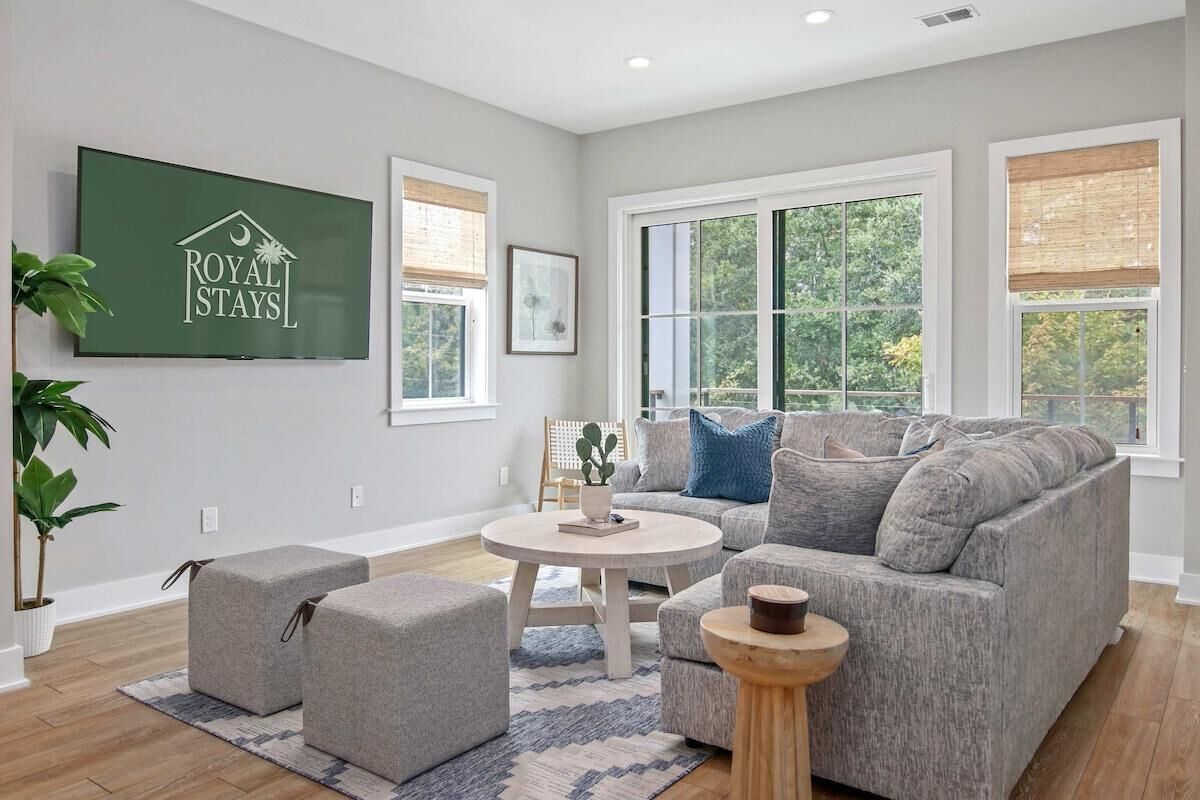 Un salon moderne avec un canapé gris, des poufs, une table basse ronde et un téléviseur affichant le logo « ROYAL STAYS ».