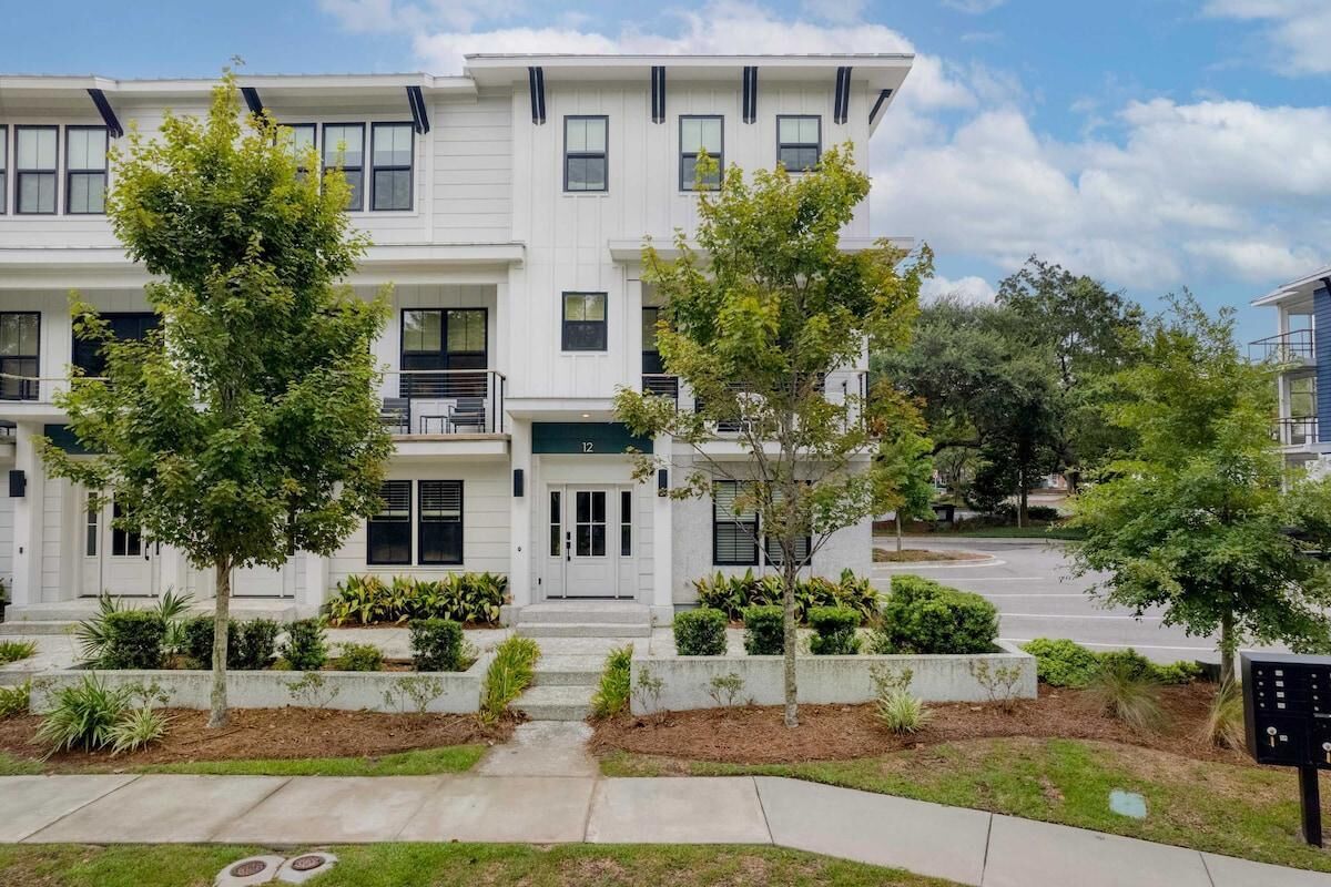 A multi-story residential building with a white exterior, multiple balconies, and a landscaped front yard with trees and shrubs.
