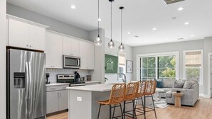 A modern kitchen with stainless steel appliances, a central island with bar stools, and a seating area with a grey sofa and a small round table. - Sincerely Shem- 7 min to Beach and Downtown CHS (Mount Pleasant)