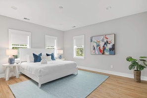 Een moderne slaapkamer met een wit bed, blauwe kussens, een blauw vloerkleed, een groot abstract schilderij en een potplant.