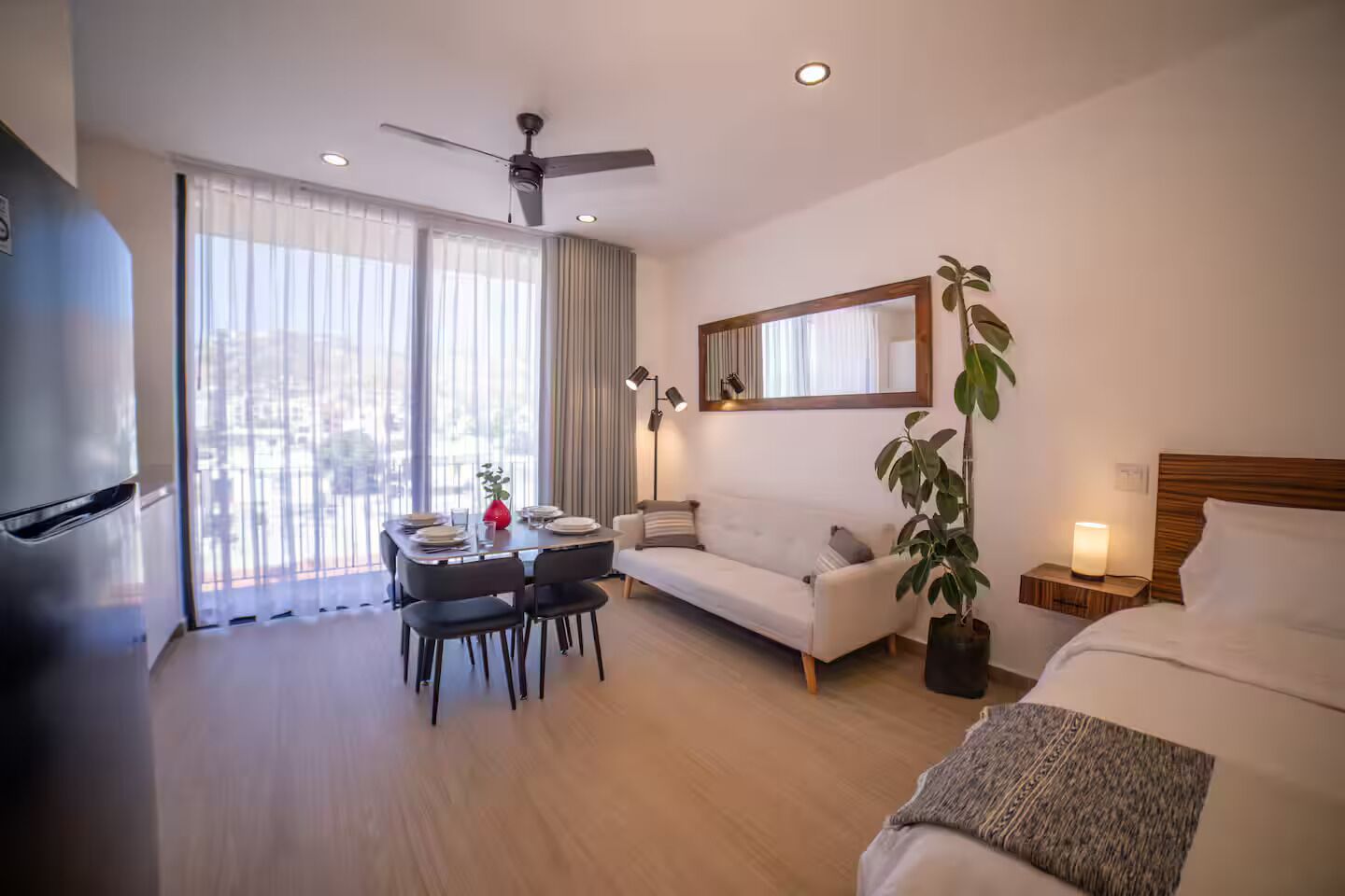 Une chambre d’hôtel comprenant un lit, un canapé, une table à manger avec des chaises, un ventilateur de plafond, une grande fenêtre avec des rideaux et une plante en pot.