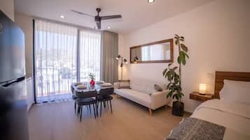 Une chambre d’hôtel comprenant un lit, un canapé, une table à manger avec des chaises, un ventilateur de plafond, une grande fenêtre avec des rideaux et une plante en pot.