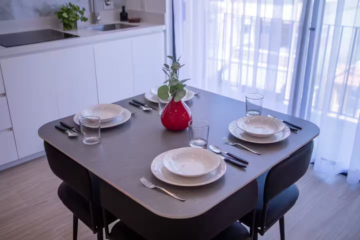 Une table à manger dressée avec des assiettes, des couverts et des verres, et un vase rouge placé au centre.