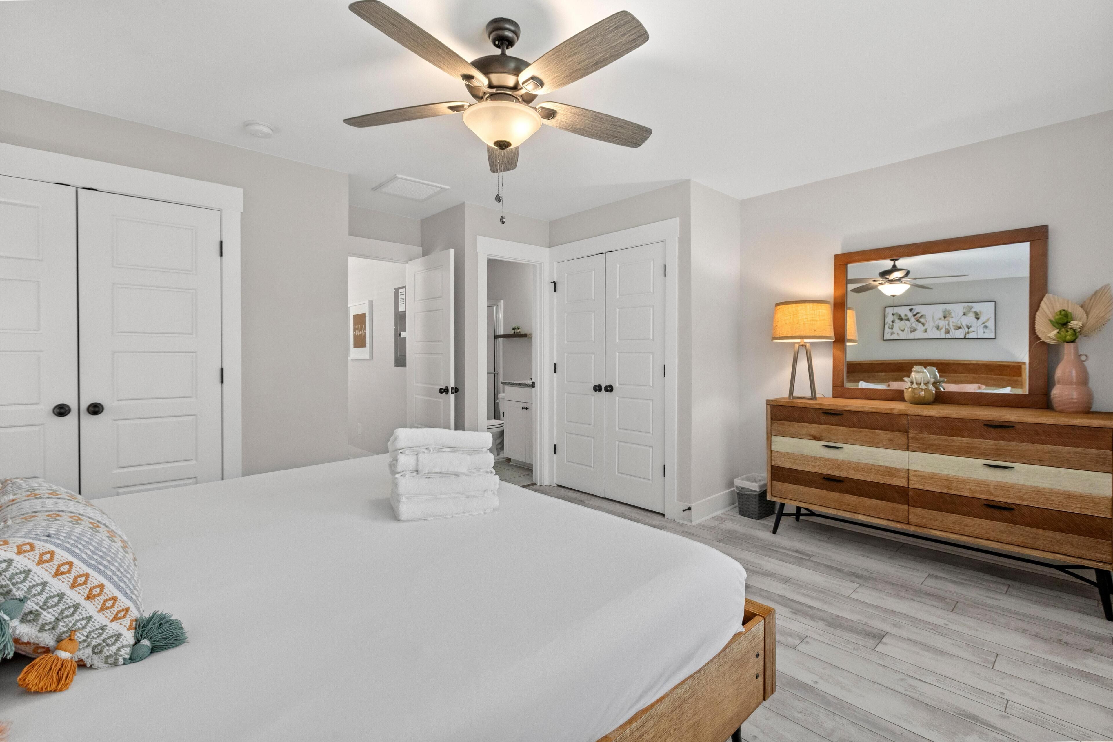 A bedroom with a large bed, a ceiling fan, white doors, a wooden dresser, and a mirror.
