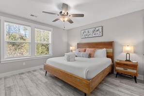 A bedroom with a wooden bed, two bedside tables with lamps, a ceiling fan, and large windows with blinds. - Creekside Bungalow-park Circle-15m Dtwn CHS (North Charleston)