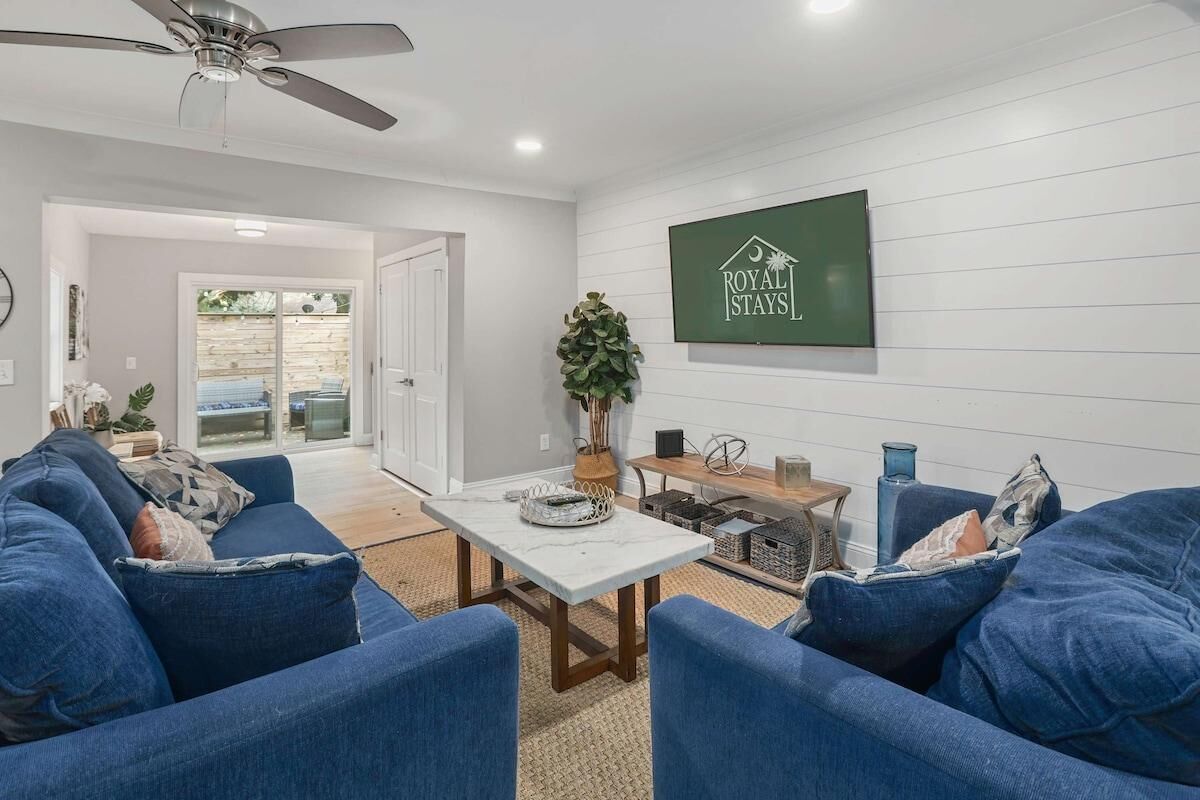 A modern living room with a blue sofa set, a white coffee table, and a ceiling fan. A sign for 'Royal Stays' is visible on the wall.