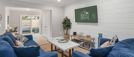 A modern living room with a blue sofa set, a white coffee table, and a ceiling fan. A sign for 'Royal Stays' is visible on the wall.