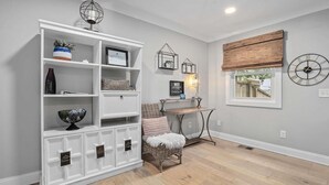 A room with a white built-in shelving unit, wicker furniture, a small table, and a window with a wooden blind. - Coastal Casa-old Village-5 Mins to Beach (Mount Pleasant)