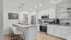 A modern kitchen with white cabinetry, a central island with bar stools, stainless steel appliances, and a chalkboard with coastal-themed drawings. - Coastal Casa-old Village-5 Mins to Beach (Mount Pleasant)