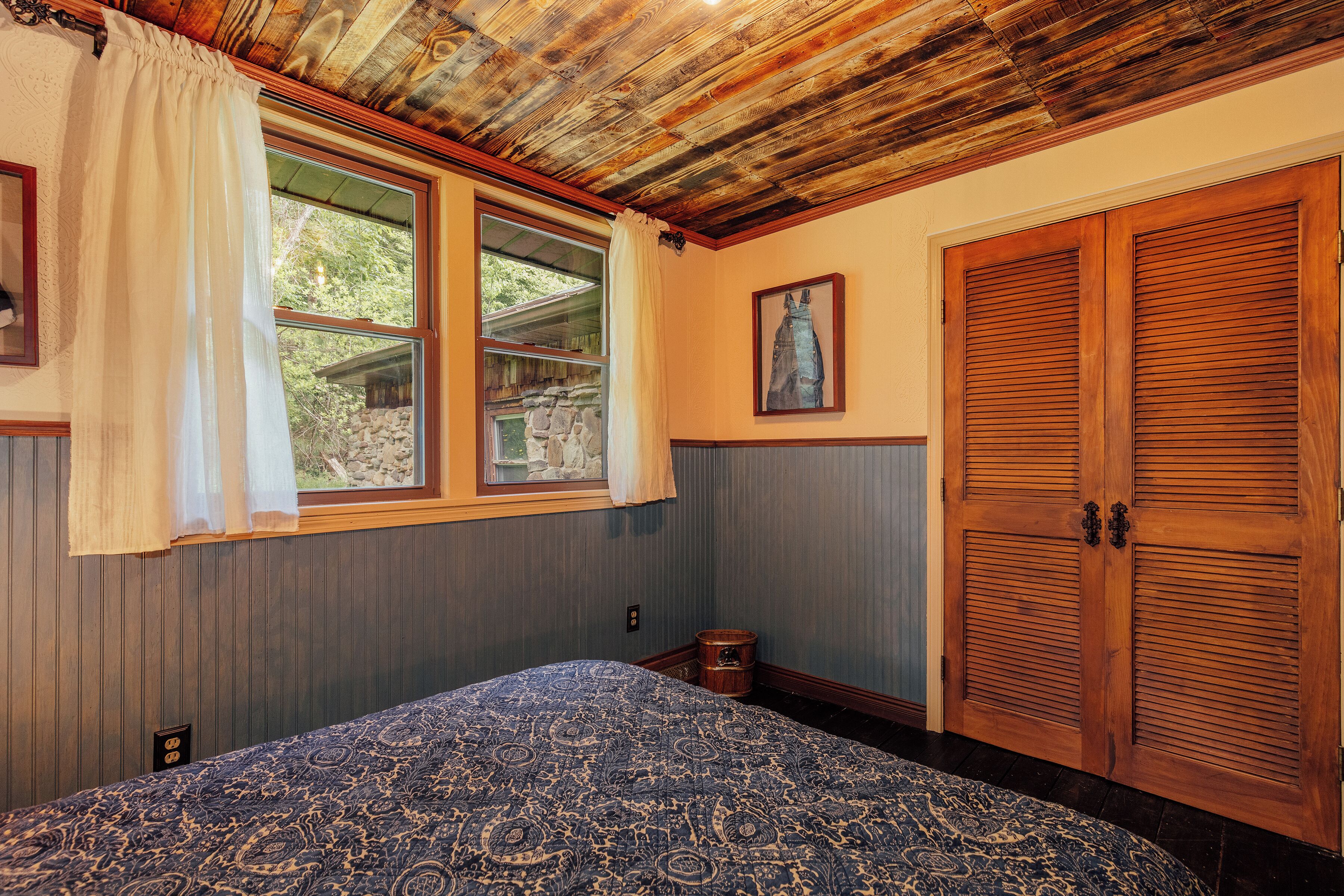 A bedroom with a bed, wooden shutters, a window with curtains, and a small chest of drawers.