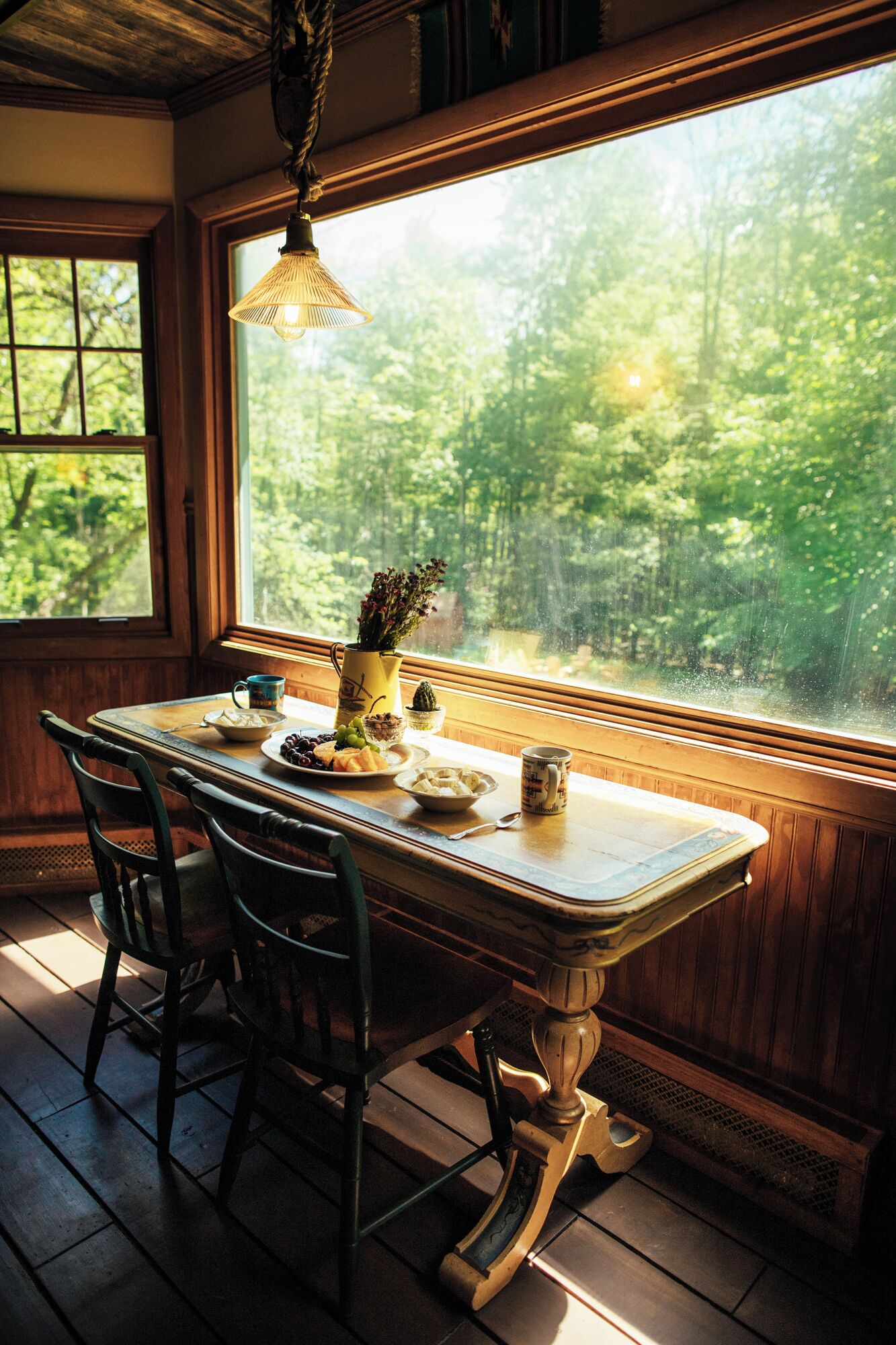 A wooden dining table with chairs, a hanging light fixture, and a view of a forest through a window.