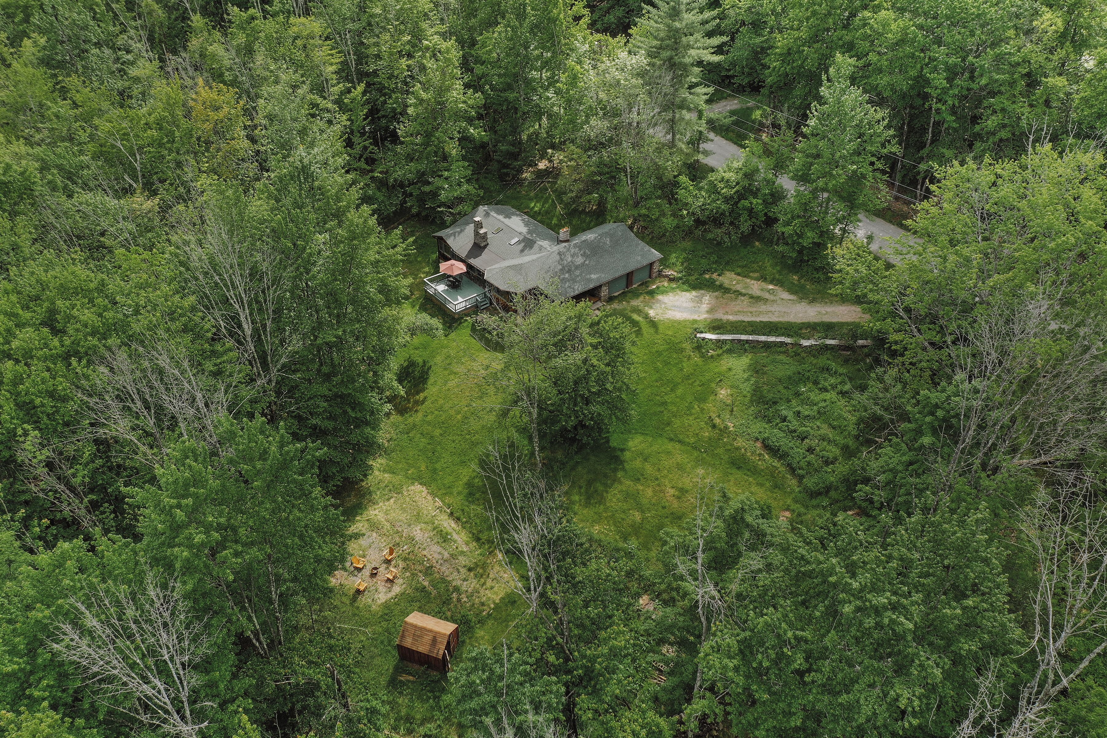 Vista aérea de una casa rodeada de un bosque denso, con un camino de entrada visible y un cobertizo pequeño.