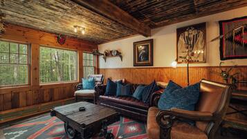 A cozy cabin interior with a leather sofa, a wooden coffee table, and a large window offering a view of the forest.
