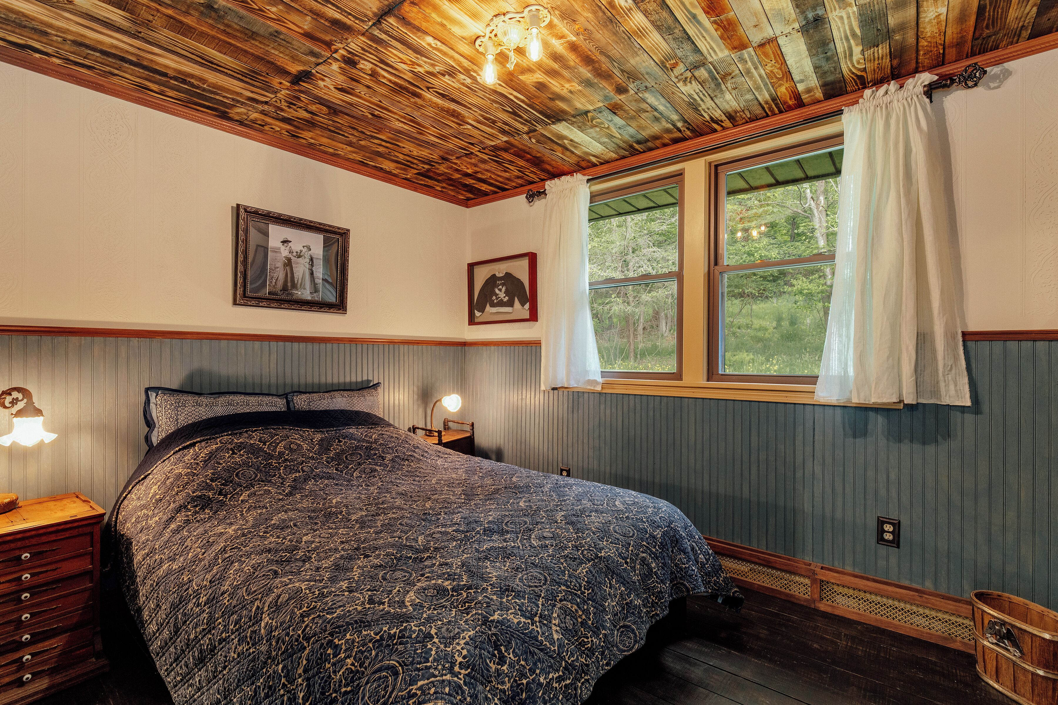 A bedroom with a large bed, a window with a view of greenery, and two framed pictures on the wall.