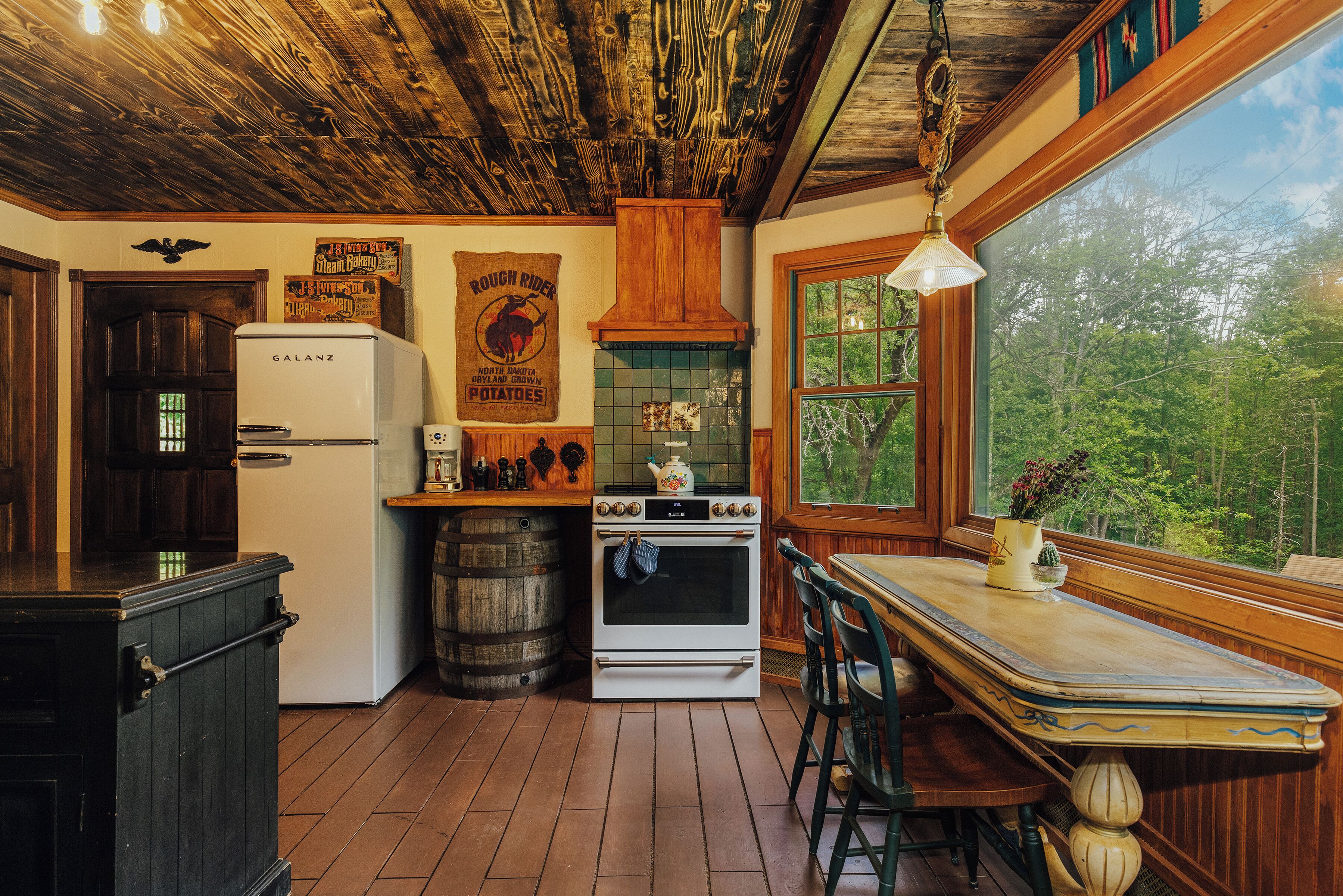 A rustic kitchen with wooden floors, a Galanz refrigerator, a stove, and a large window overlooking greenery.