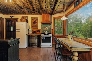 A rustic kitchen with wooden floors, a Galanz refrigerator, a stove, and a large window overlooking greenery.