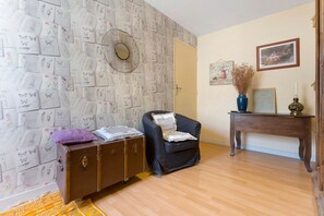 A room with a patterned wallpaper, a vintage suitcase, a black armchair, a wooden side table with a vase, and a framed picture on the wall.