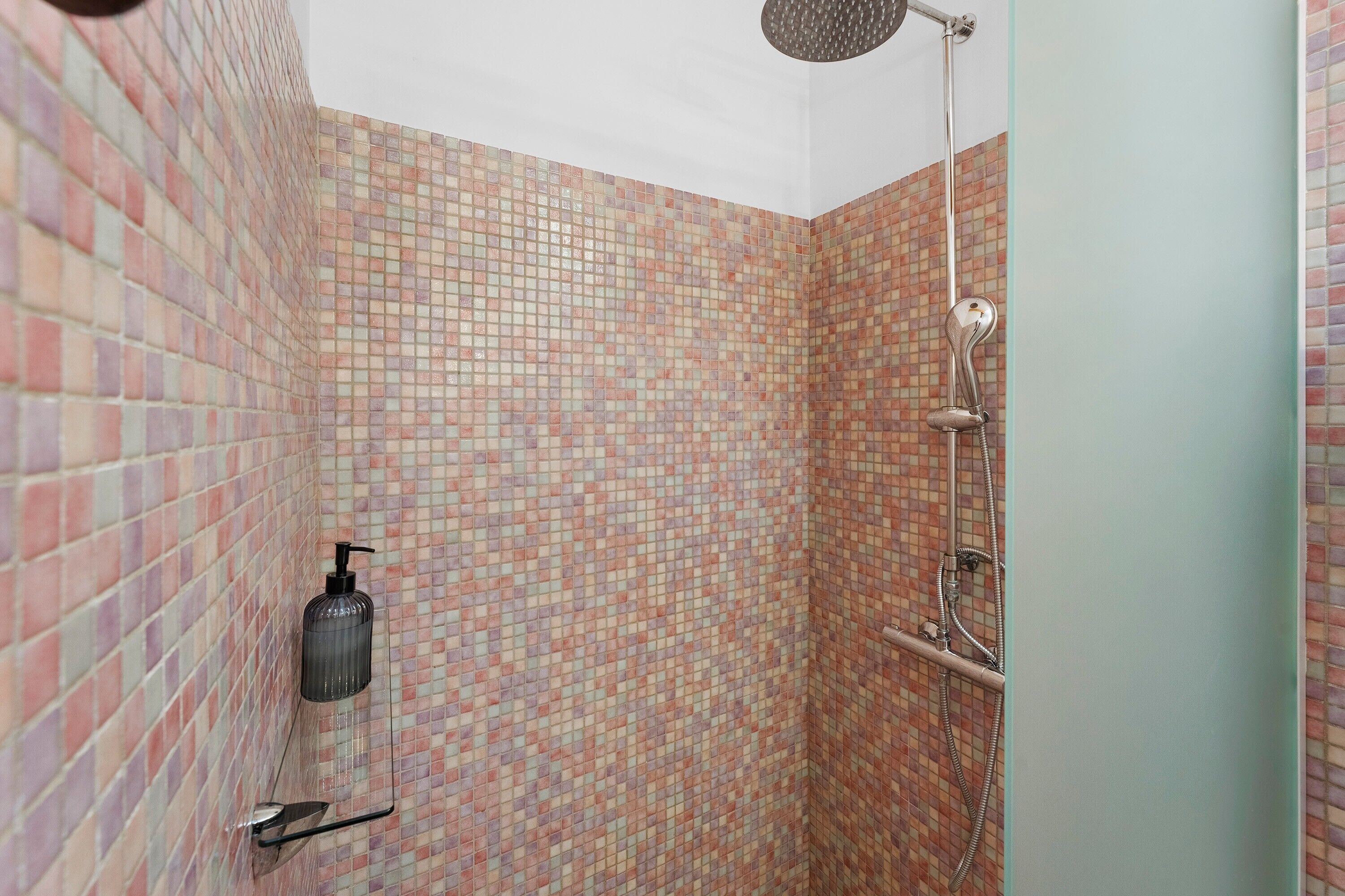 Zona de ducha con pared de azulejos, cabezal de ducha de mano y una botella de champú.