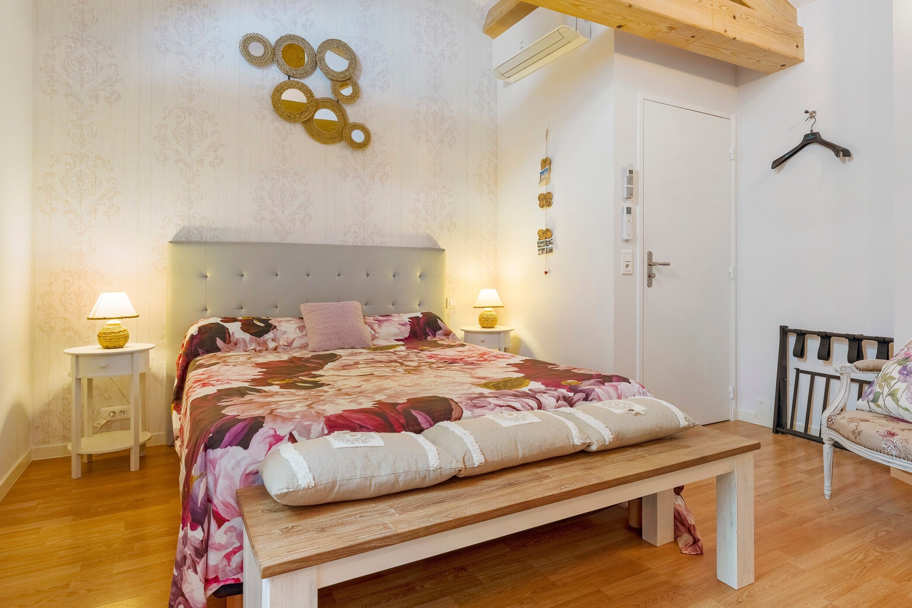 Une chambre à coucher comprenant un lit, des tables de chevet, un banc, un luminaire suspendu et une porte.