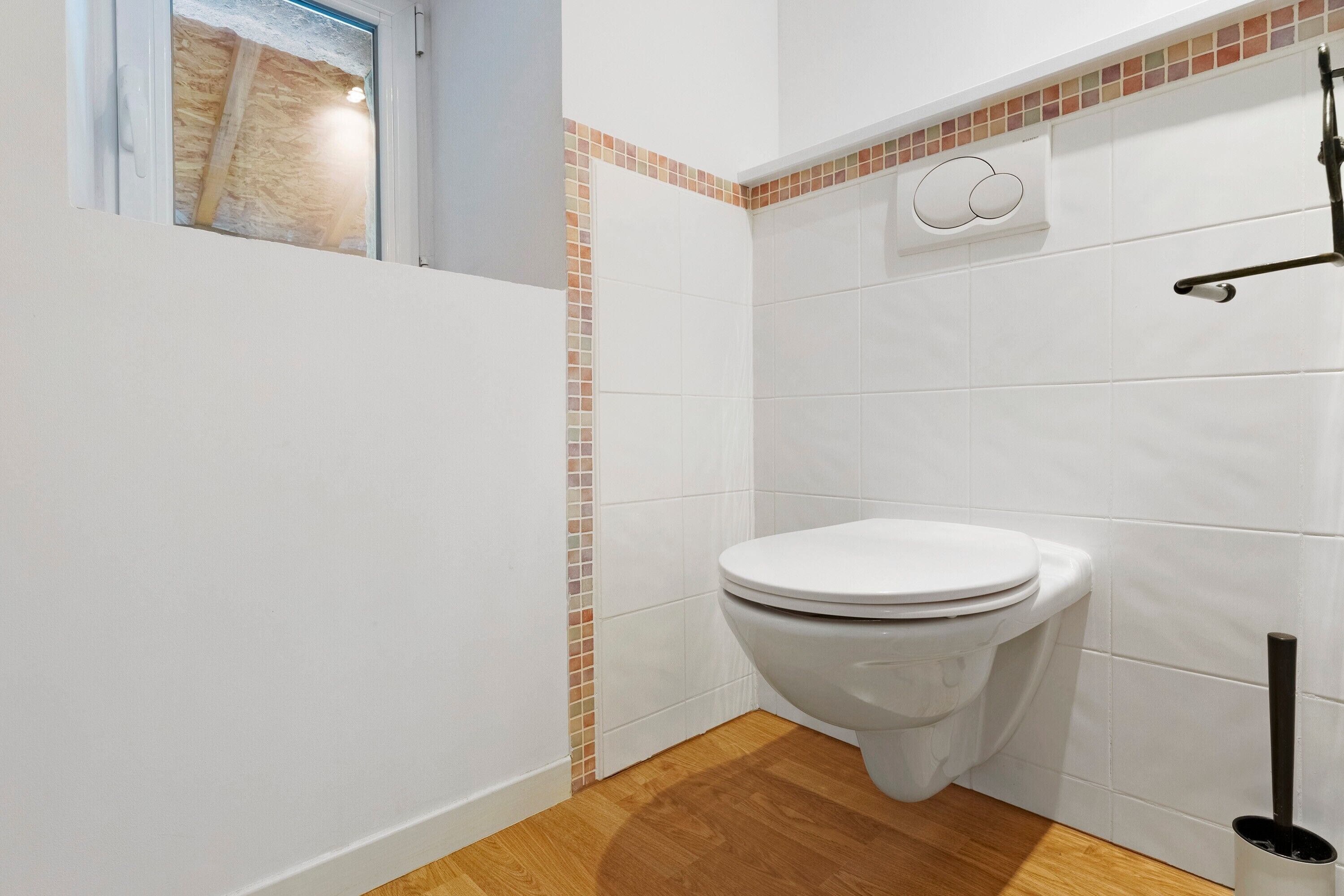 Une petite salle de bain avec des toilettes blanches, un plancher en bois et un mur carrelé avec une petite fenêtre.