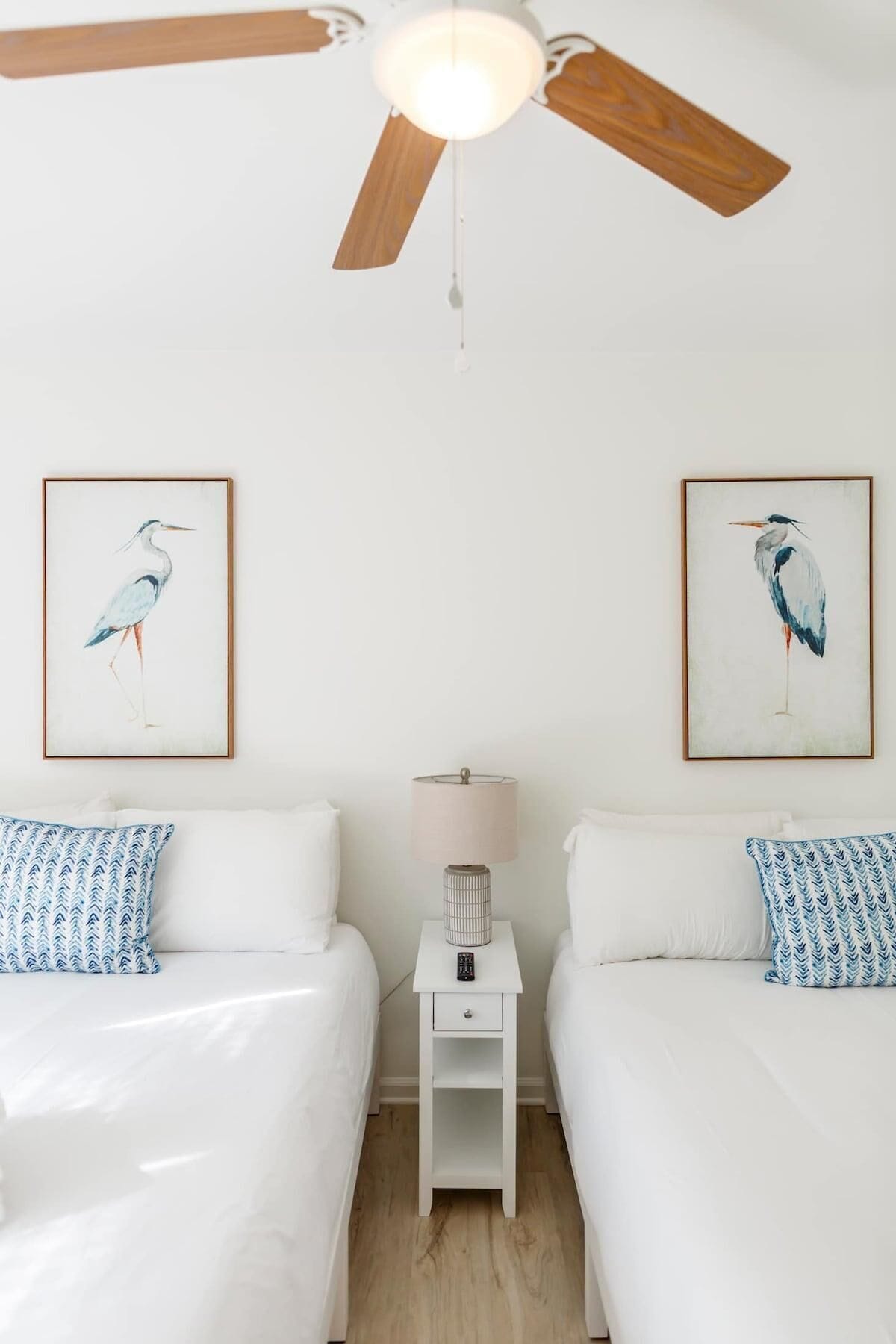 A bedroom with two beds, a ceiling fan, a nightstand with a lamp, and two framed bird paintings on the wall.