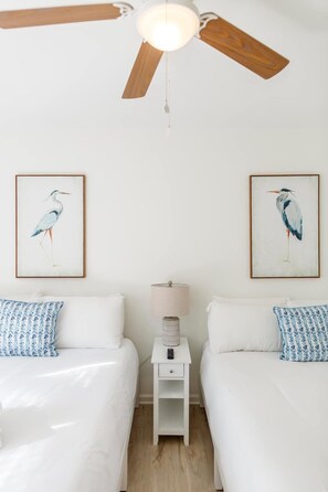 Un dormitorio con dos camas, un ventilador de techo, una mesita de noche con lámpara y dos cuadros enmarcados con pájaros en la pared.