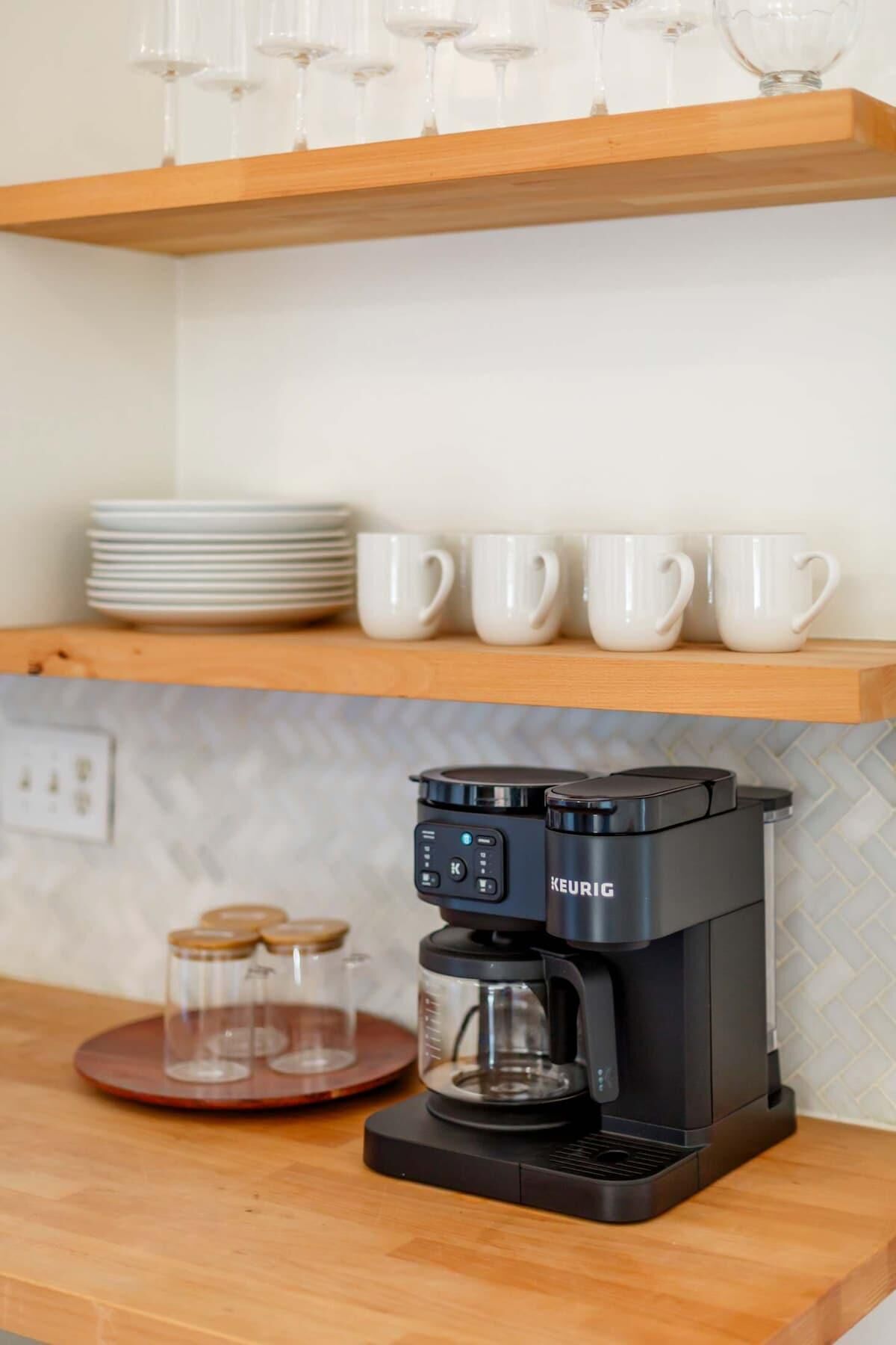 Eine Küche mit einer Kaffeemaschine auf der Arbeitsfläche, einem Regal mit Tellern und Tassen sowie einer Wand mit Zickzackmuster.