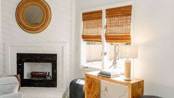A room with a fireplace, a round mirror, two windows with blinds, a table with a lamp, and a sofa.
